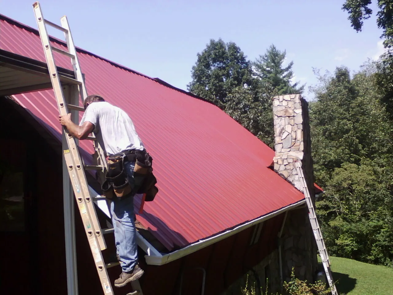 Roofer on a ladder beside a red metal roof and stone chimney — Soffit & Fascia Repair by Roofing Brothers in Morgan